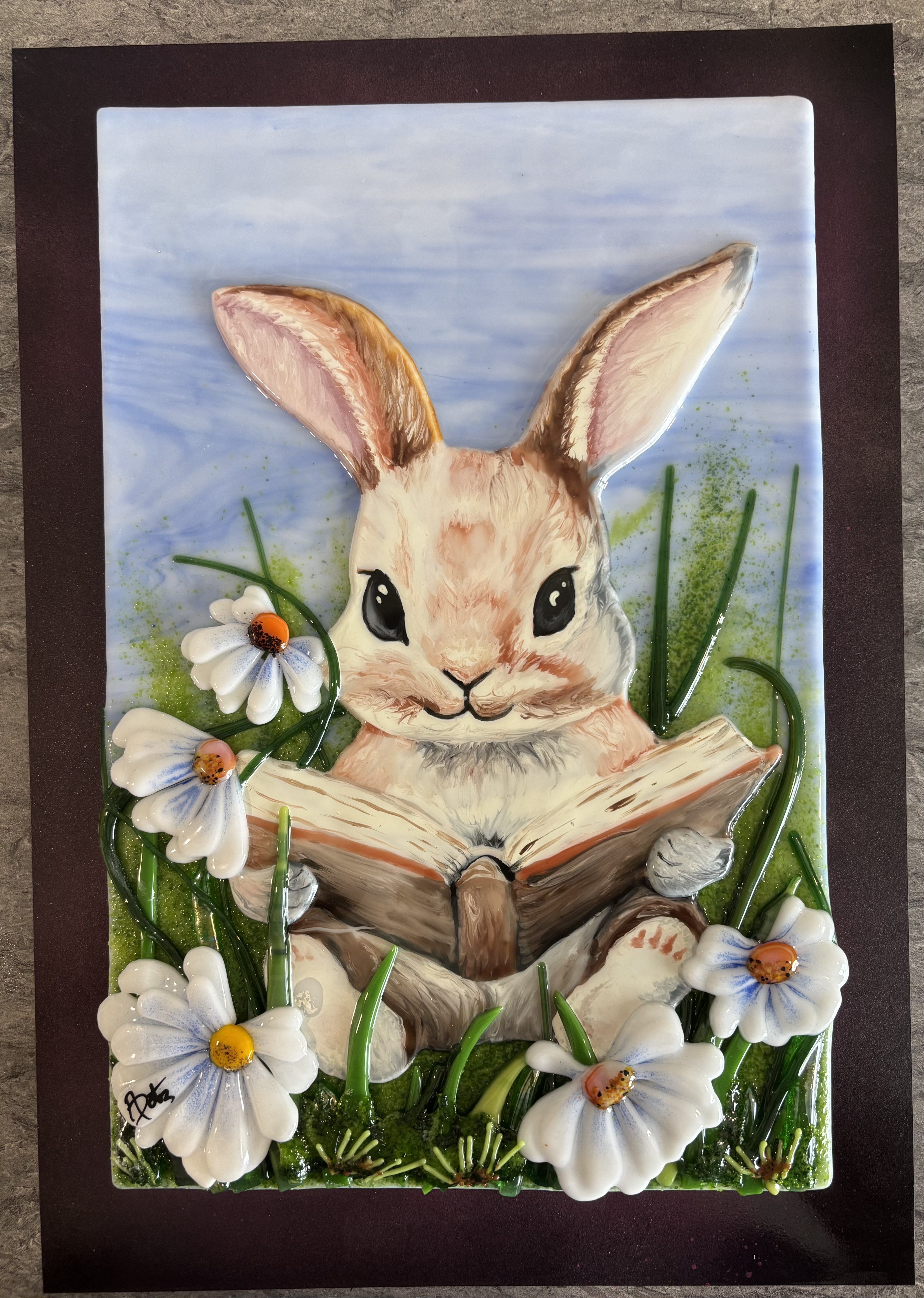 Whimsical fused-glass bunny reading a book among daisies.