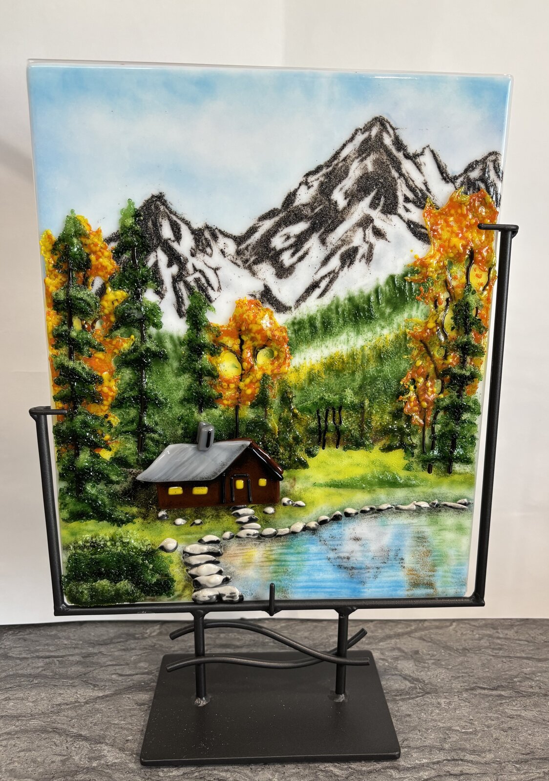 A fused-glass panel of a cabin by a pond beneath snow-capped mountains and evergreens, on a black metal stand.