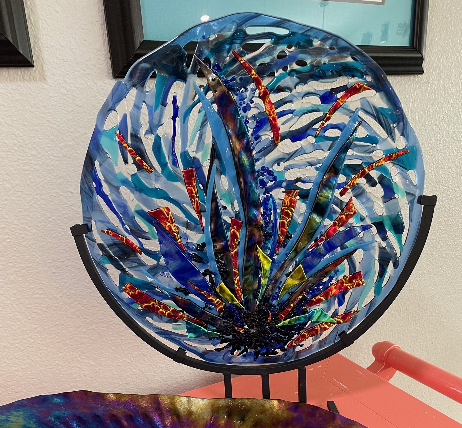 Circular Blue: a round fused-glass platter in deep blue with white, red, and gold abstract strokes, mounted on a black metal stand.