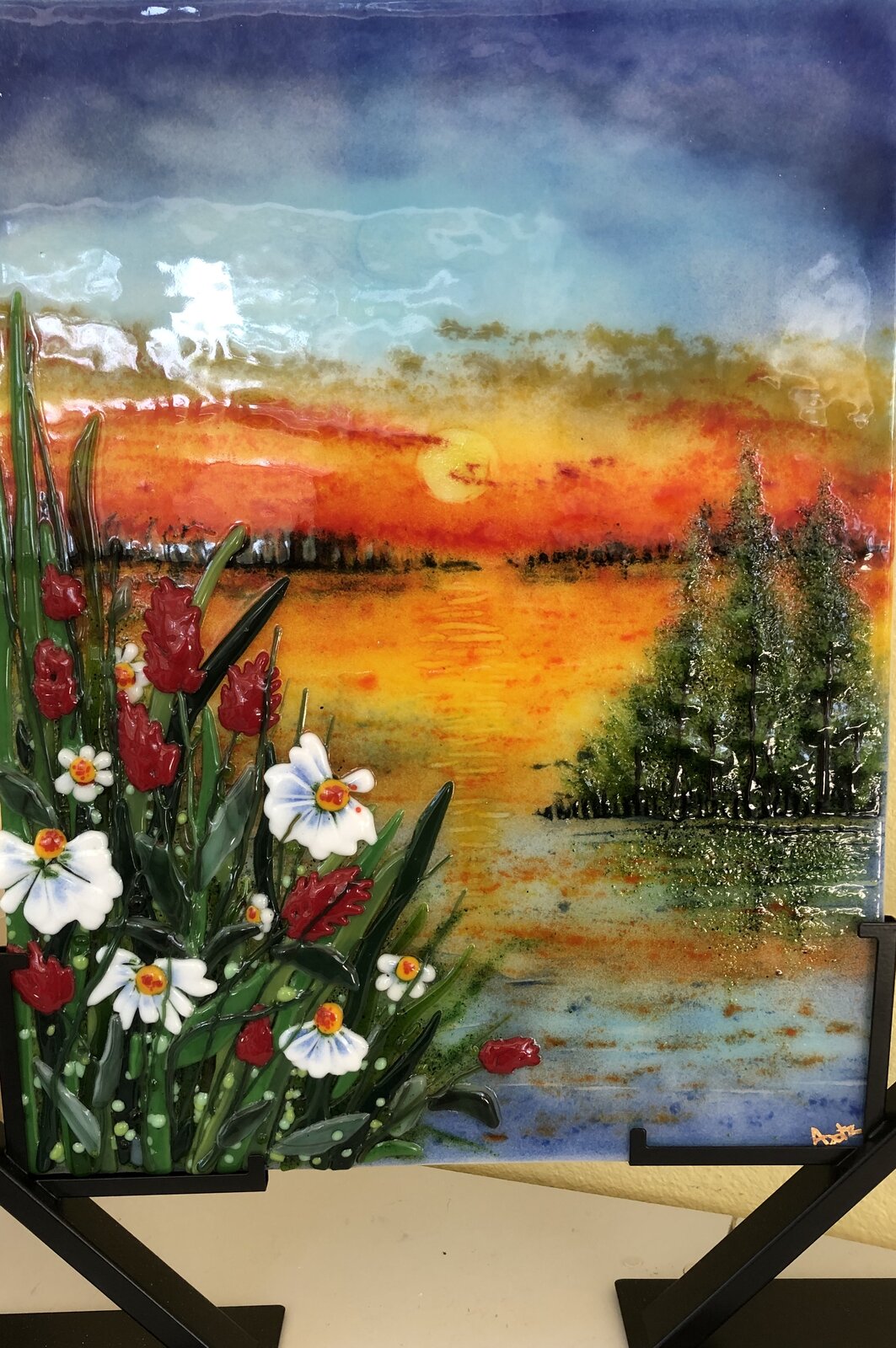 Vertical fused-glass panel of a sunset over a lake, orange sky above blue water, evergreens on the far shore and sculpted daisies and red wildflowers in the foreground, on a black metal stand.