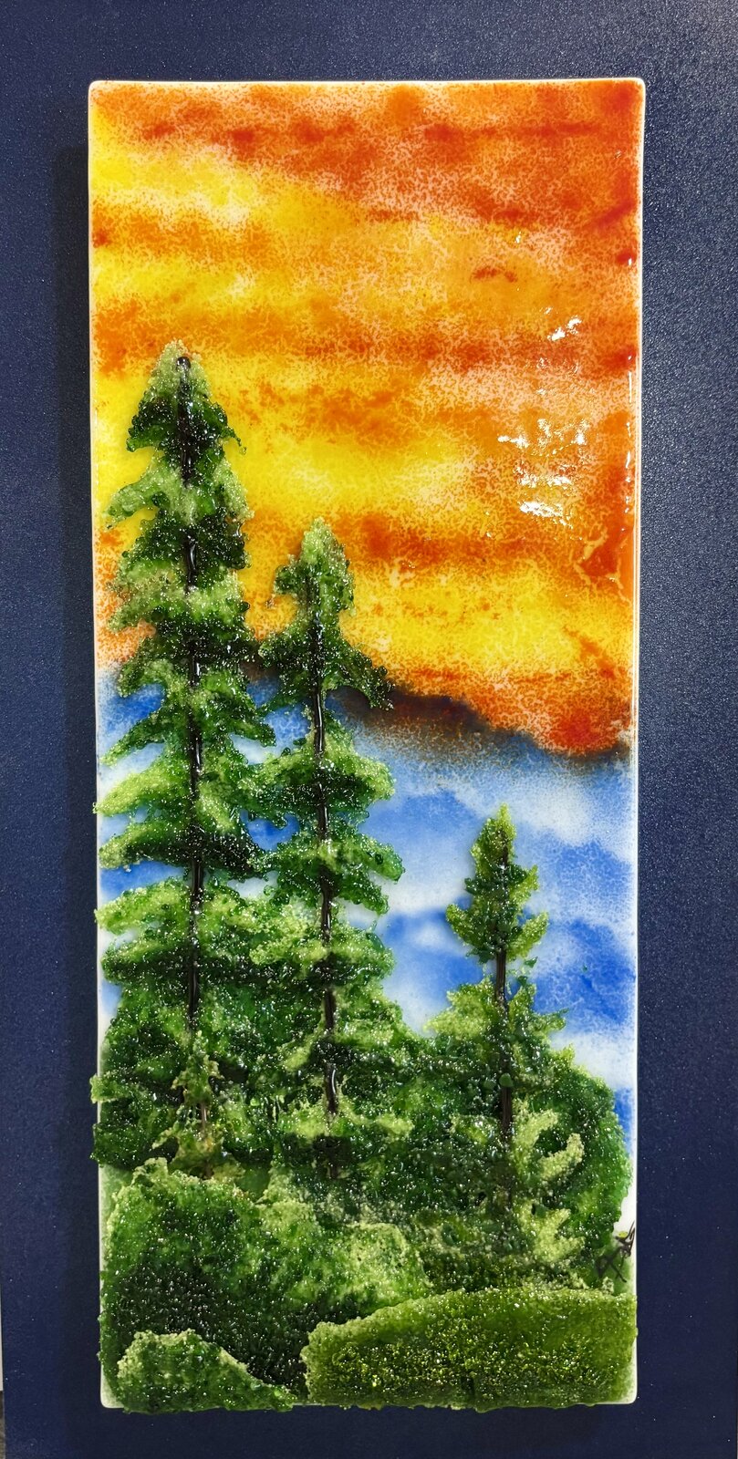 A tall fused-glass panel of evergreens against an orange and gold sunset sky.
