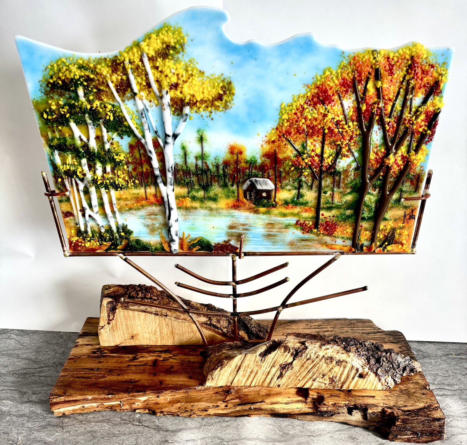 A fused-glass fall landscape of birches and red maples around a pond, mounted above a live-edge wood base.