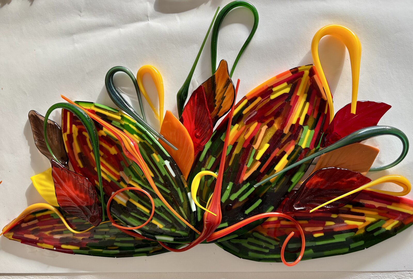 Sculptural fused-glass fan of striped autumn leaves in green, red, orange, and yellow, with hand-pulled glass loops curling above.