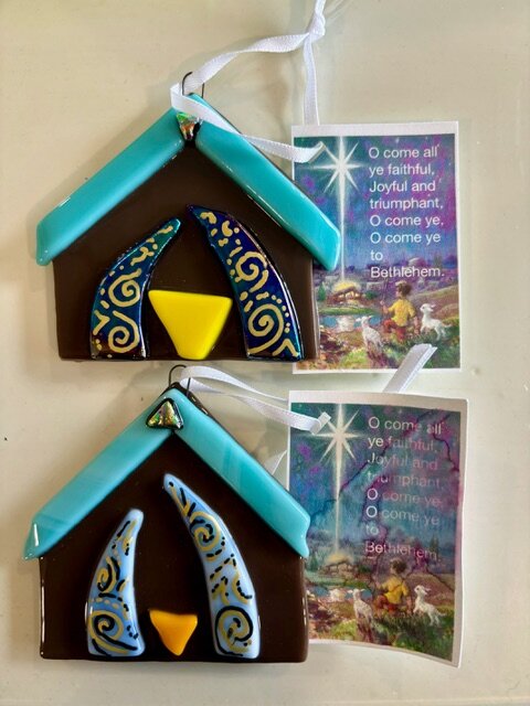 Nativity: fused-glass stable ornaments with a teal roof and patterned blue glass figures leaning over the child.