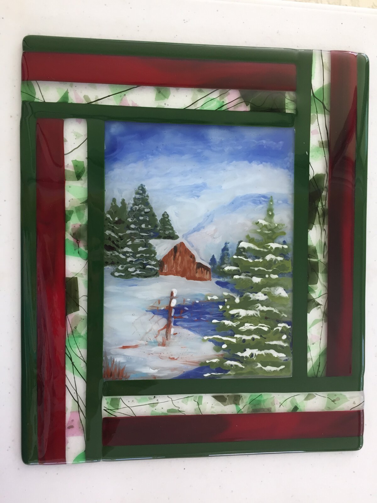 Fused-glass panel of a snowy cabin beside a creek with snow-covered evergreens, set in a red and green glass border like a quilted frame.