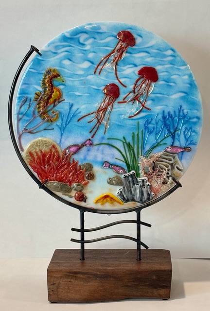 Underwater Fantasy: a circular fused-glass panel of luminous coral, sea fans, and bubbles in turquoise and blue, mounted on a sculptural metal stand.